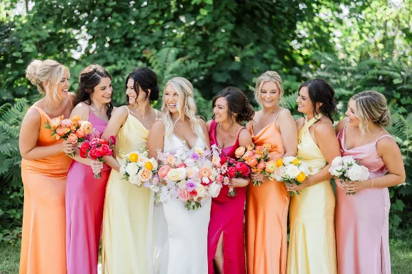 A bride in a white gown surrounded by bridesmaids in colorful dresses holding bouquets outdoors.