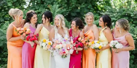 A bride in a white gown surrounded by bridesmaids in colorful dresses holding bouquets outdoors.