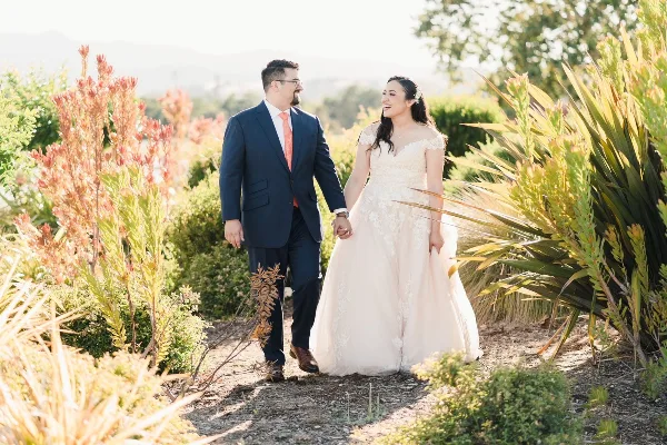 A bride and groom walking hand-in-hand through a garden with lush greenery and flowers.
