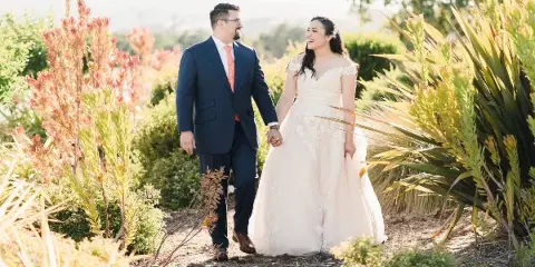 A bride and groom walking hand-in-hand through a garden with lush greenery and flowers.