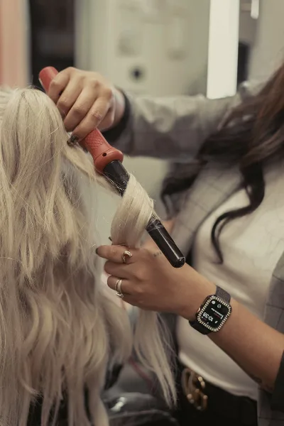A close-up of a hairstylist using a curling iron to style a client's long blonde hair.