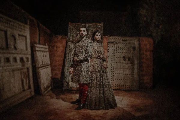 A couple in traditional wedding attire standing together in a dimly lit rustic setting with carved wooden panels.