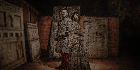 A couple in traditional wedding attire standing together in a dimly lit rustic setting with carved wooden panels.