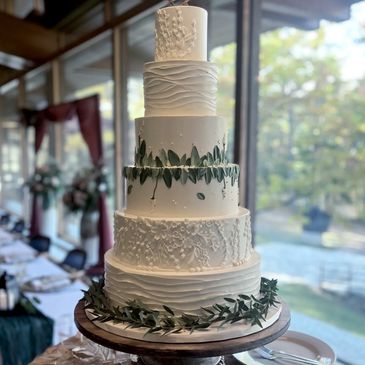 A five-tier white wedding cake decorated with green leafy garlands, displayed on a wooden stand in a bright room.