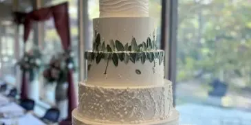 A five-tier white wedding cake decorated with green leafy garlands, displayed on a wooden stand in a bright room.
