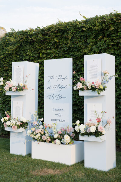 A custom white wedding signage installation with floral arrangements and text reading 'The People who Helped Us Bloom' and 'Deanna & Dimitrios'.