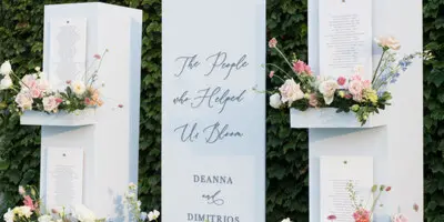 A custom white wedding signage installation with floral arrangements and text reading 'The People who Helped Us Bloom' and 'Deanna & Dimitrios'.