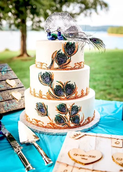 A three-tier white wedding cake decorated with peacock feathers and figurines, sitting on a table with a cake server and a wooden sign.