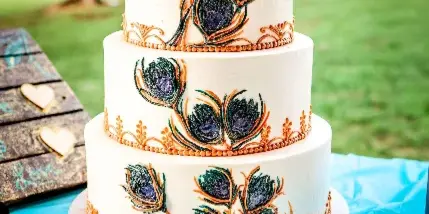 A three-tier white wedding cake decorated with peacock feathers and figurines, sitting on a table with a cake server and a wooden sign.