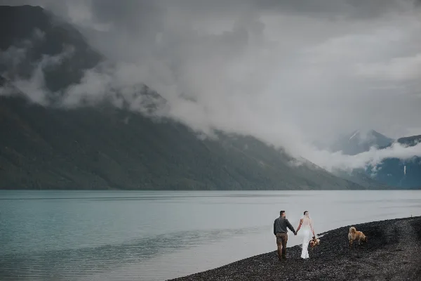 A bride and groom holding hands while walking along a rocky shoreline next to a large lake with mountains in the background.