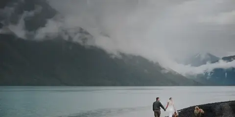 A bride and groom holding hands while walking along a rocky shoreline next to a large lake with mountains in the background.