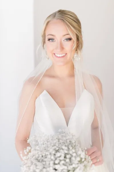 Bride in a white wedding gown and veil holding a bouquet of white flowers, smiling with styled hair and makeup.