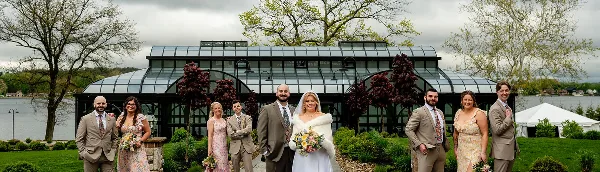 A wedding party poses in front of a glass-roofed conservatory structure with a lake in the background.