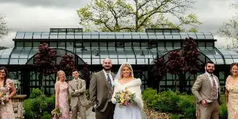 A wedding party poses in front of a glass-roofed conservatory structure with a lake in the background.