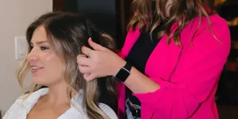 A hairstylist in a pink blazer styling the hair of a seated client in a white top.