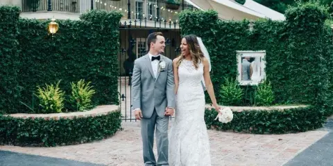 A bride and groom standing together in a courtyard with manicured hedges and a building entrance.