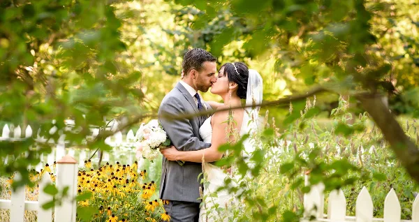 A bride and groom sharing a kiss in a garden setting with a white picket fence and yellow flowers.