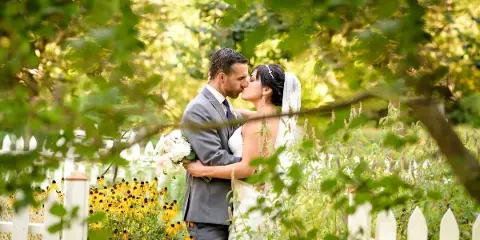 A bride and groom sharing a kiss in a garden setting with a white picket fence and yellow flowers.
