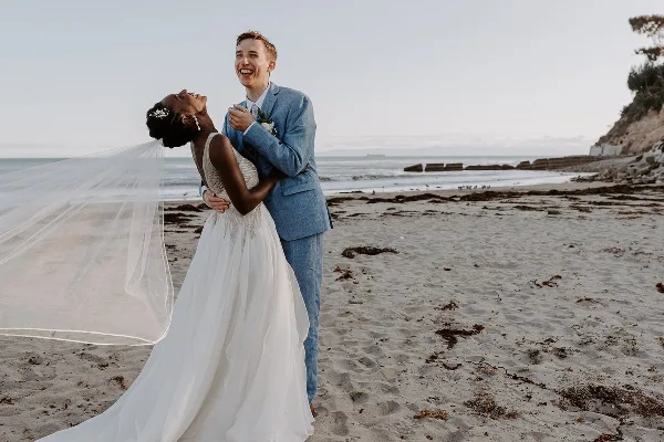 A bride and groom laughing together on a sandy beach, with the bride's veil blowing in the wind.