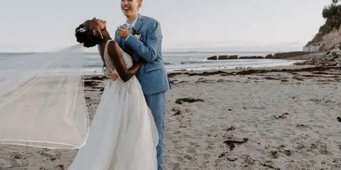 A bride and groom laughing together on a sandy beach, with the bride's veil blowing in the wind.