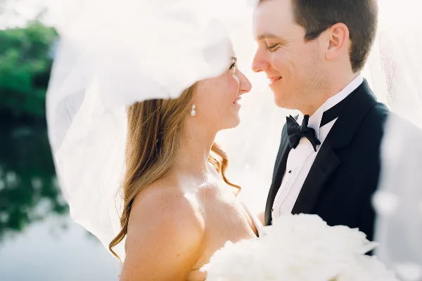A close-up photograph of a bride and groom smiling at each other, with the bride wearing a veil and the groom in a tuxedo.