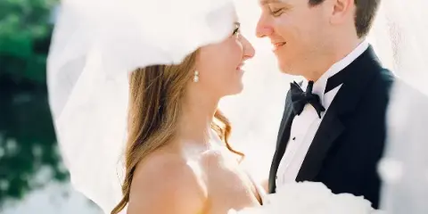 A close-up photograph of a bride and groom smiling at each other, with the bride wearing a veil and the groom in a tuxedo.