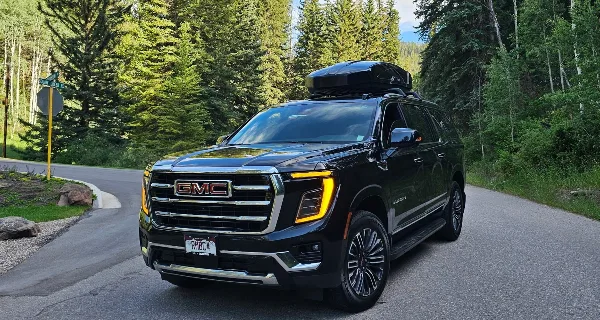 A black GMC Yukon SUV with a roof cargo box parked on a paved road surrounded by evergreen trees.