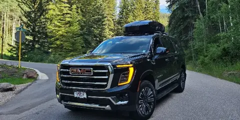 A black GMC Yukon SUV with a roof cargo box parked on a paved road surrounded by evergreen trees.