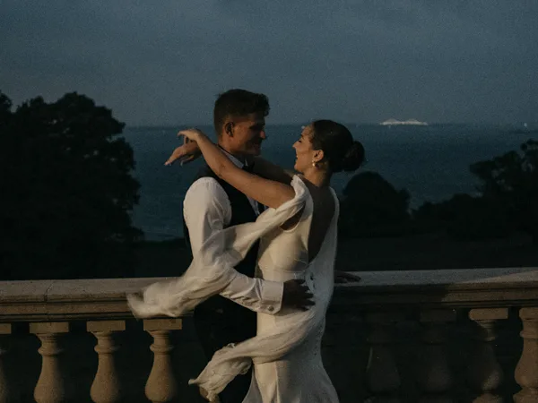 A bride and groom dancing on a stone balcony overlooking the ocean at dusk, captured in a film photography style.