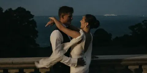 A bride and groom dancing on a stone balcony overlooking the ocean at dusk, captured in a film photography style.