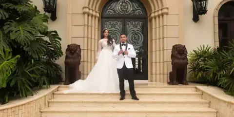 A bride in a white gown and a groom in a tuxedo standing on the steps of a grand building entrance.