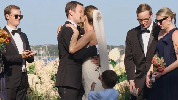 A cinematic still from a wedding film showing a bride and groom sharing a kiss during an outdoor ceremony, surrounded by wedding party members and floral arrangements.