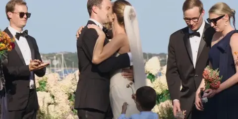 A cinematic still from a wedding film showing a bride and groom sharing a kiss during an outdoor ceremony, surrounded by wedding party members and floral arrangements.