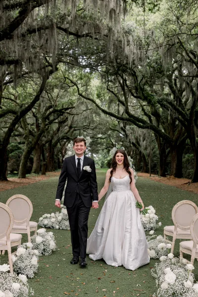 A bride and groom walking down an outdoor aisle decorated with white flowers and chairs under a canopy of trees.