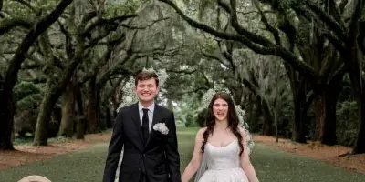 A bride and groom walking down an outdoor aisle decorated with white flowers and chairs under a canopy of trees.