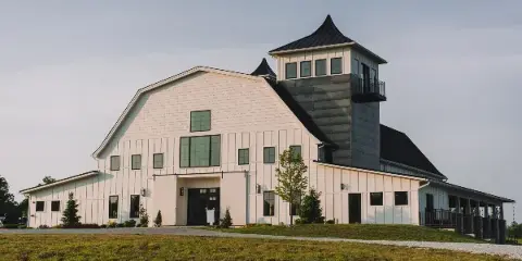 Exterior view of a large white barn-style building with a black metal tower and cupola, set on a grassy lawn.