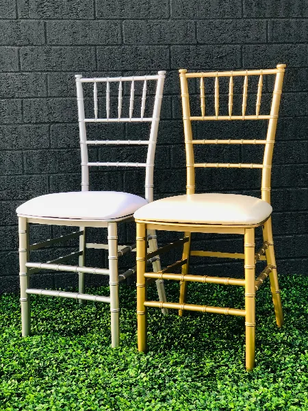 Two Chiavari chairs, one white and one gold, with cushioned seats positioned on a green surface against a dark brick wall.