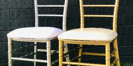 Two Chiavari chairs, one white and one gold, with cushioned seats positioned on a green surface against a dark brick wall.