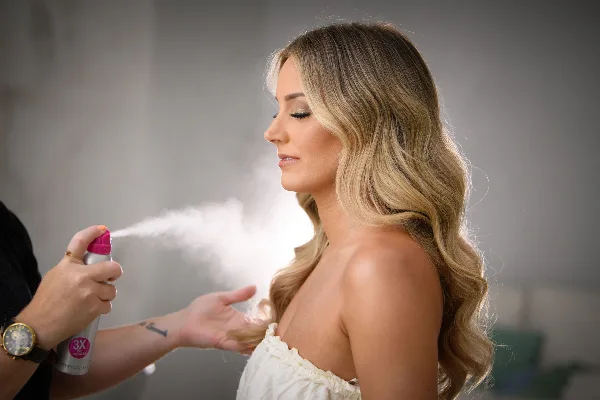 A stylist sprays a finishing product onto a bride's long, wavy blonde hair.