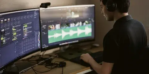 A video editor wearing headphones working at a desk with two monitors displaying video editing timelines and footage.