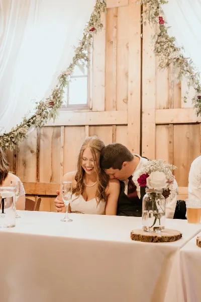 A bride and groom seated at a reception table with floral arrangements and rustic wooden decor.