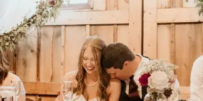 A bride and groom seated at a reception table with floral arrangements and rustic wooden decor.