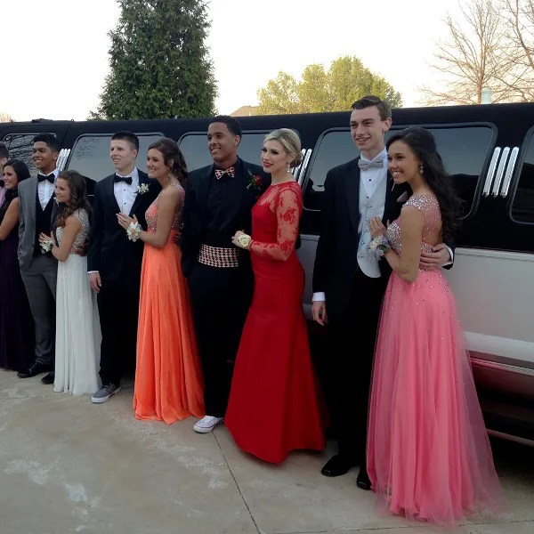 A group of formally dressed young adults posing with a black limousine.