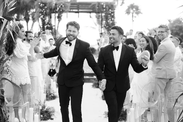 Black and white photograph of a smiling couple in formal wear walking down a wedding aisle holding hands, with guests clapping on either side.