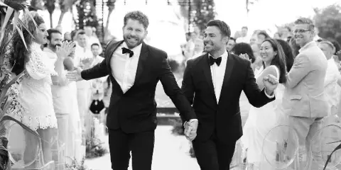 Black and white photograph of a smiling couple in formal wear walking down a wedding aisle holding hands, with guests clapping on either side.