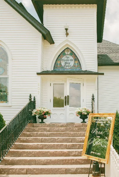 Exterior view of a white Victorian-style event venue with stone steps, floral arrangements, and a wedding welcome sign.
