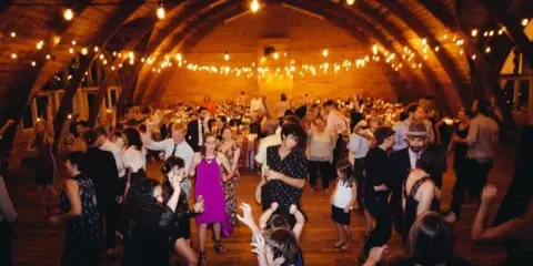 A crowded wedding reception with guests dancing in a rustic wooden barn venue illuminated by string lights.