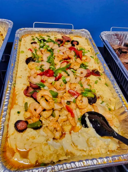 A large aluminum catering tray containing a prepared dish of shrimp, sausage, and rice with a black serving spoon.