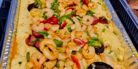A large aluminum catering tray containing a prepared dish of shrimp, sausage, and rice with a black serving spoon.