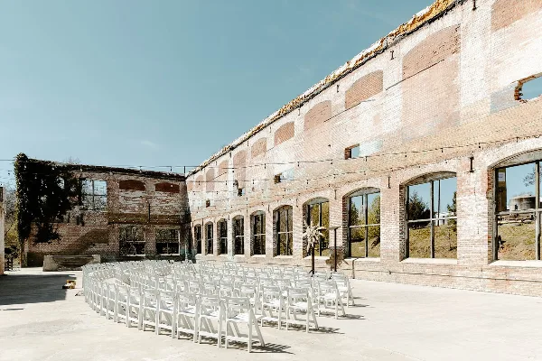 Rows of white chairs set up for a wedding ceremony in front of a historic brick industrial mill building.